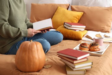 Evde yatakta çörekler ve sonbahar yapraklarıyla kitap okuyan bir kadın.