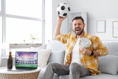 Futbol topu ve parayla evdeki kanepede oturan mutlu genç adam. Spor bahsi konsepti