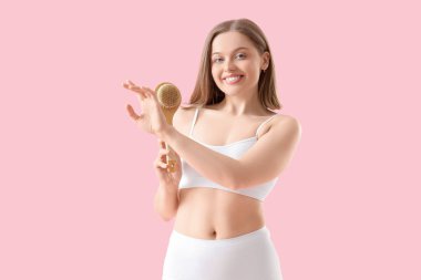 İç çamaşırlı genç bir kadın pembe arka planda fırçayla koluna masaj yapıyor.