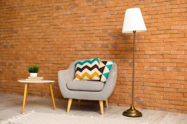 Yastıklı gri koltuk, sehpa ve kahverengi tuğla duvarın yanında standart lamba.