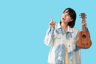 Ukuleleli güzel genç bir kadın mavi arka plandaki bir şeyi işaret ediyor.