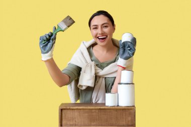 Fırça ve konserve kutularıyla genç bir kadın sarı arka plana ahşap masa boyuyor.