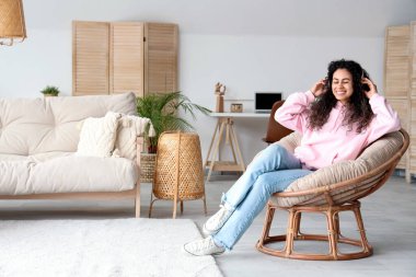 Kulaklıklı genç Afro-Amerikalı kadın evdeki koltukta dinleniyor.