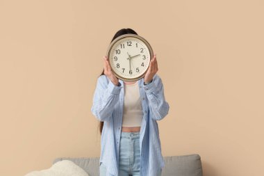 Oturma odasında bej duvarın yanında saati tutan genç bir kadın.