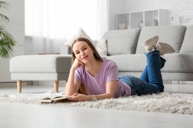 Genç ve güzel bir kadın şık bir oturma odasında kitapla yerde yatıyor.