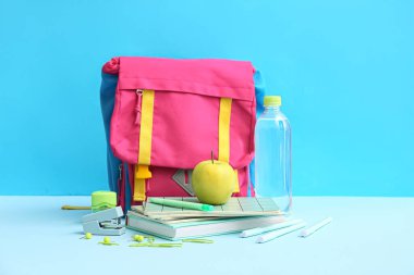 Farklı kırtasiye malzemeleriyle renkli okul çantası, mavi arka planda bir şişe su ve elma.