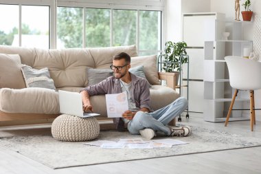 Gözlüklü genç işadamı ofiste yerde doküman ve bilgisayarla çalışıyor.