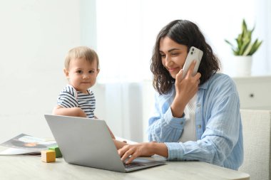 Evde cep telefonuyla konuşurken dizüstü bilgisayarla çalışan tatlı bir bebek ve genç anne.