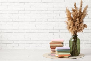 Beyaz tuğla duvarın yanında otlu vazo ve kitap yığınları.