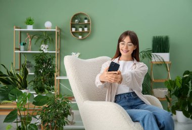 Genç bayan bahçıvan cep telefonuyla selfie çekiyor evdeki ev bitkilerinin yanındaki koltukta.