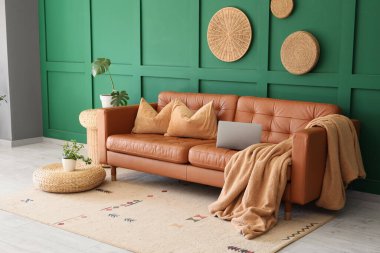 Yeşil duvarın yanındaki şık deri koltukta hasır koltuklar, ev bitkileri ve dizüstü bilgisayarla oturma odasının içinde.