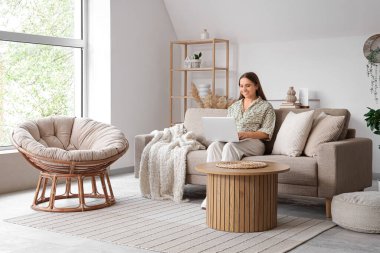 Güzel genç bayan şık oturma odasındaki rahat gri koltukta dizüstü bilgisayar kullanıyor.