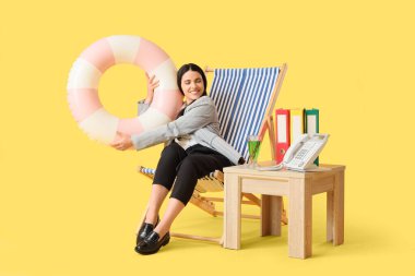 Güverte sandalyesinde yüzme yüzüğü ve sarı arka planda masası olan genç bir iş kadını. Yaz tatili kavramı