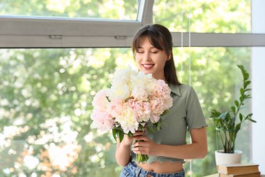 Evinin penceresinin yanında şakayık çiçekleri olan gülümseyen genç bir kadın.