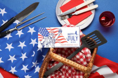 ABD bayrağı, 4 Temmuz 'da Mutlu Yıllar yazılı tebrik kartı, piknik sepeti ve mavi arka planda barbekü aletleri. Bağımsızlık Günü kutlamaları