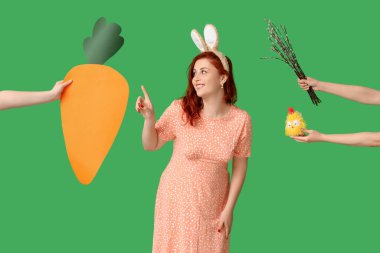 Beautiful young woman in bunny ears pointing at hands with paper carrot, pussy willow branches and toy chick on green background. Easter celebration