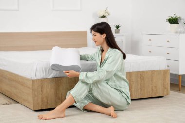 Pijamalı, ortopedik yastıklı, yatak odasında yerde oturan genç bir kadın.