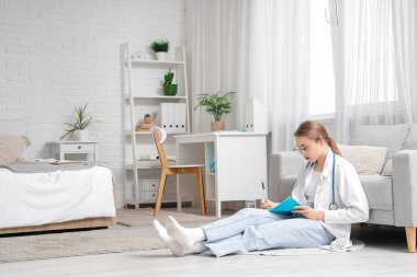 Bayan tıp öğrencisi evde yerde defterle ders çalışıyor.