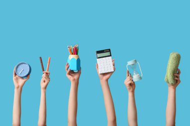 Mavi zemin üzerinde kırtasiye, hesap makinesi ve alarm saati olan birçok el