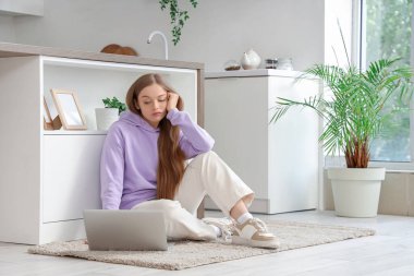 Mutfakta yerde modern dizüstü bilgisayar kullanan üzgün genç bir kadın.