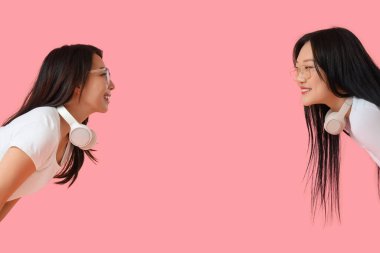 Güzel, mutlu, kulaklıklı Asyalı kız kardeşler pembe arka planda eğleniyorlar.