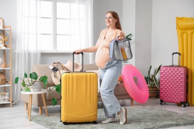 Seyahat aksesuarları ve bavuluyla evde genç hamile bir kadın.