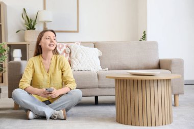 Oturma odasındaki gri kanepenin yanında cep telefonu kullanan gülümseyen genç kadın.