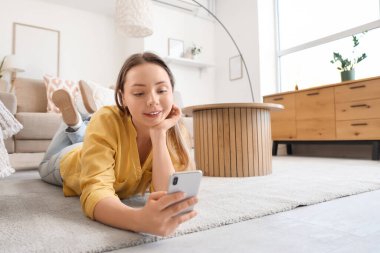 Oturma odasında cep telefonu kullanan gülümseyen genç bir kadın.