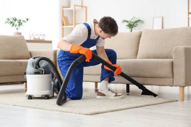 Modern elektrikli süpürgeyle oturma odasında halıyı temizleyen genç bir erkek hademe.