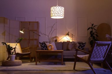 Lüks oturma odasının içinde rahat gri kanepe, koltuklar, sehpa ve geceleri parlayan lambalar.