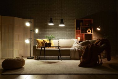 Şık oturma odasının içinde rahat gri kanepe, koltuk, sehpa ve akşam lambaları parlıyor.