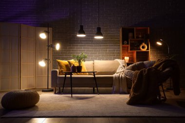 Şık oturma odasının içinde rahat gri kanepe, koltuk, sehpa ve akşam lambaları parlıyor.