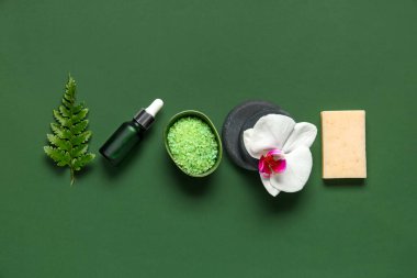 Yeşil arka planda bir kase deniz tuzu, banyo malzemeleri ve bir şişe kozmetik ürünle kompozisyon