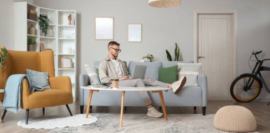 Gözlüklü genç adam evdeki kanepede dizüstü bilgisayar kullanıyor.