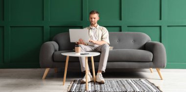 Genç sakallı adam evdeki kanepede dizüstü bilgisayar kullanıyor.