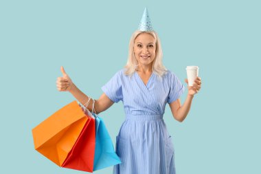 Parti şapkalı olgun bir kadın, elinde kahve fincanı ve mavi arka planda baş parmağını gösteren alışveriş torbalarıyla.