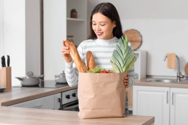Mutfaktaki masada içi taze yiyecek dolu alışveriş çantasıyla genç bir Asyalı kadın.