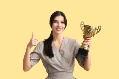 Altın bardaklı genç kozmetikçi sarı arka planda baş parmak gösteriyor.