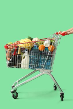 Yeşil arka planda farklı yiyeceklerle dolu alışveriş arabasını iten kadın.