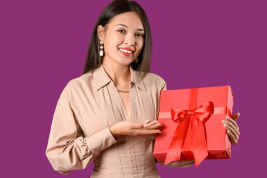 Güzel genç Asyalı kadın mor arka plandaki hediye kutusunu işaret ediyor.