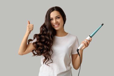 Gri arka planda baş parmağını gösteren saç maşası olan güzel genç bir kadın.