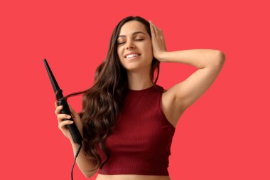 Beautiful young woman with curling iron on red background