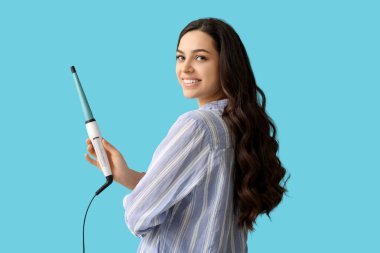 Beautiful young woman with curling iron on blue background