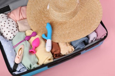 İçinde giysiler, hasır şapka ve pembe arka planda seks oyuncakları olan bavulu aç.