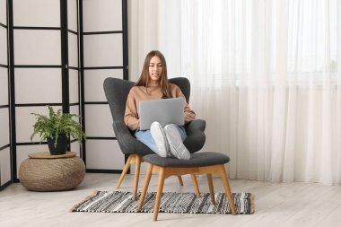 Evinin penceresinin yanındaki gri rahat koltuklu dizüstü bilgisayarı olan güzel genç bir kadın.