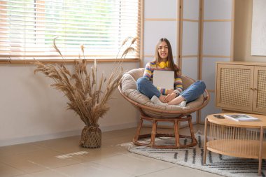 Odadaki koltukta otururken dizüstü bilgisayar kullanan genç bir kadın.