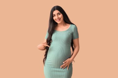 Genç hamile bir kadın kahverengi arka planda karnını işaret ediyor.