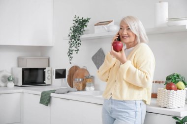Elma tutan ve mutfakta cep telefonuyla konuşan olgun bir kadın.