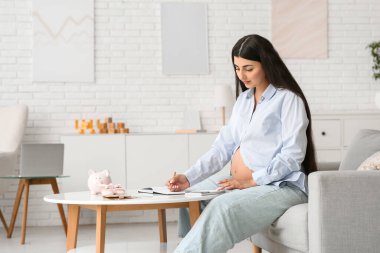 Genç hamile kadın evdeki kanepede hesap makinesi kullanıyor. Annelik Faydası konsepti