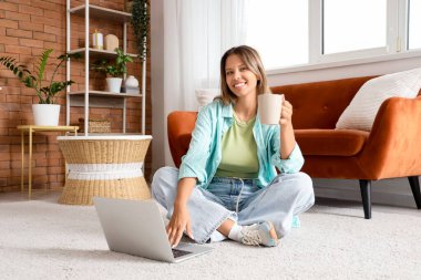 Evde dizüstü bilgisayar kullanan çay içen genç bir kadın.